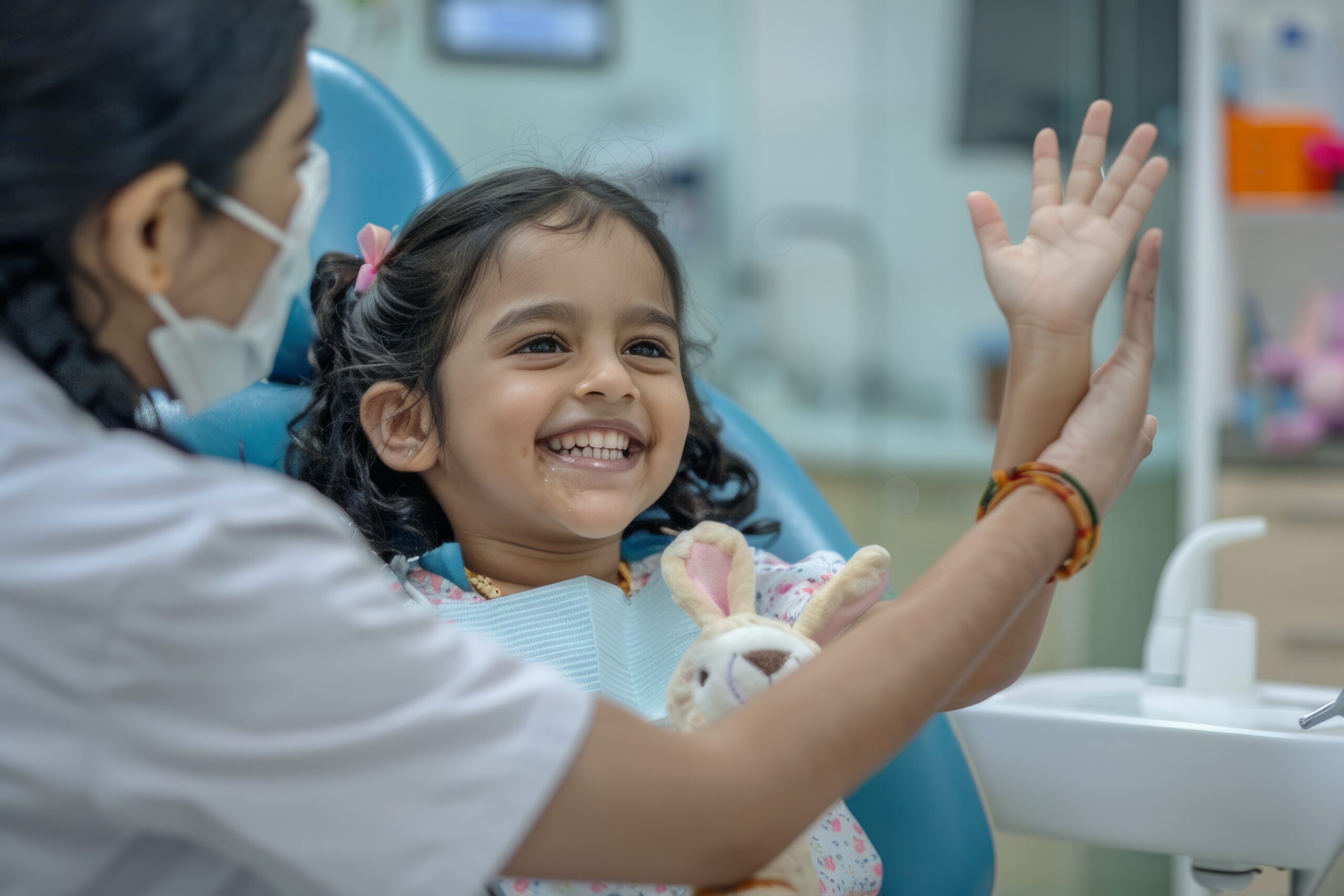 An Indian girl, holding a toy rabbit, sits in the dental chair and cheerfully gives a high-five to the nurse during the doctor's appointment.
